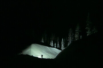 A lone skier walks at night up a snow trail illuminated by the headlamp the snow and trees are...