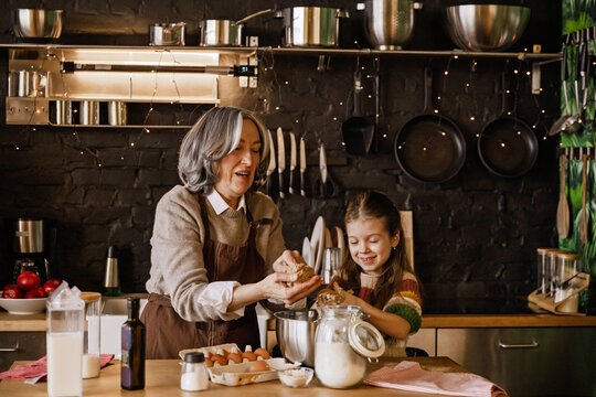 A White grandmother and granddaughter bake cookies in a cozy kitchen, mixing dough, shaping treats, and laughing together, surrounded by a warm light and some festive decor, creating holiday memories.
