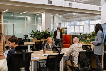 A White elder man is interacting with a group of colleagues in a modern and spacious office space, while discussing ideas and working together in a collaborative and professional environment.