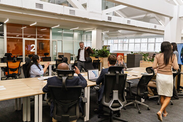 Obraz premium A White elder man enters a modern office, greeted by a multinational group of colleagues in business attire, engaging in a discussion with laptops and documents around them in a workspace.
