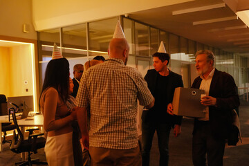 A multinational group of office workers celebrates a White elder man's retirement in a warmly lit work space, wearing party hats, sharing drinks, exchanging gifts, and enjoying a gathering together.
