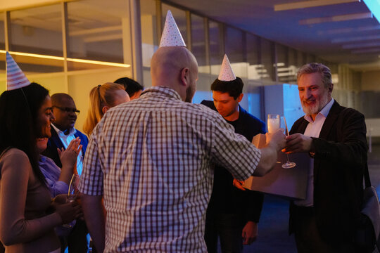 A middle-aged man in a blazer receives a farewell gift and a champagne toast from colleagues at an office party. Diverse group of men and women wearing party hats are gathered, celebrating together.
