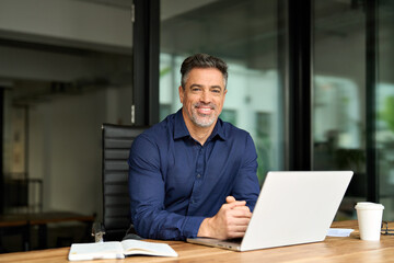 Portrait of happy business man leader of middle age working on laptop in office. Senior executive...