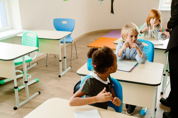 Obraz premium A group of children sit at their desks in a bright classroom. A blonde boy in a light blue shirt holds a pencil while looking focused, while a Black girl in uniform is engaged in an activity.