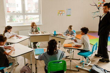 A group of young students sits at their desks in a classroom, drawing and writing with colored pencils under the supervision of their teacher. 