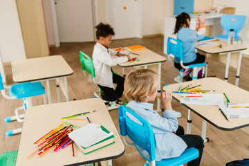 Obraz premium boy in a light blue shirt sits at his desk, holding a pencil while looking at his drawing. Other students are focused on their creative activities