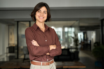 Happy confident young Hispanic professional business woman standing at work in office arms crossed...