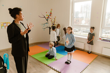 A teacher speaks to a group of students standing on colorful mats in a classroom. The diverse children listen attentively during a school lesson.