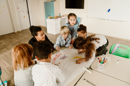 A teacher and a group of children work together on a creative drawing project in a classroom. The diverse children use markers to color a large paper, engaging in a fun and interactive learning.