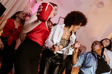 A group of young women sings and dances at a lively indoor party, holding a microphone and drinks. The diverse friends enjoy a fun night of karaoke, music, and celebration.