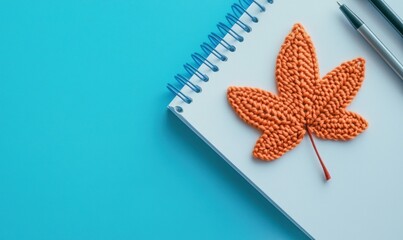 A bright blue background features an orange crochet leaf on a notebook with pens, suggesting autumn crafting.