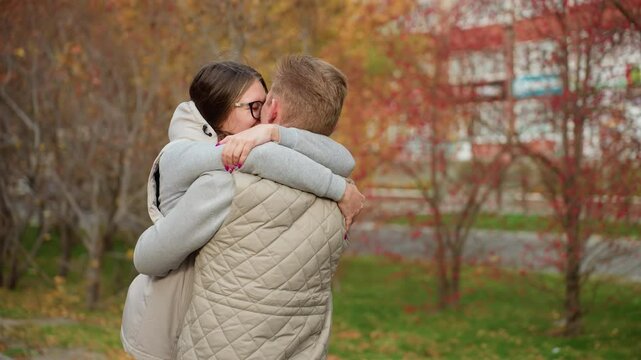 Man and wife hug each other tightly as they share warm kiss outdoors in autumn park, dressed in matching jackets, surrounded by trees with colorful leaves, enjoying romantic moment together