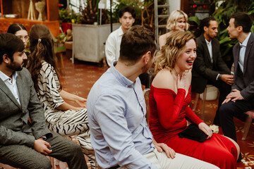 A diverse group of adults sits together at an event, smiling and laughing. One woman in a red gown is sitting with a man in a blue shirt. The group is enjoying a conversation at stylish indoor venue.