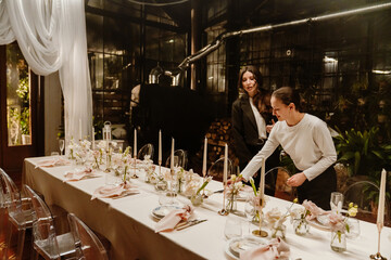 Two women arranging a decorated wedding reception table with some elegant floral centerpieces, candles, and big soft pink napkins, preparing the intimate setting for a celebration in a charming venue.