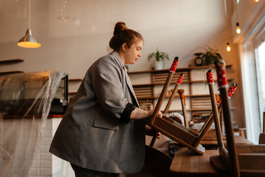 A White female cafe owner preparing for opening by arranging chairs, actively working on the renovation of her new cafe, showcasing her dedication to creating a welcoming and stylish space.