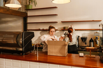 A White female cafe owner and a White female worker with red hair organizing and unpacking supplies...