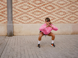 A White young woman doing squats outdoors on the pavement near a brick wall, dressed in a pink sweatshirt, shorts, and sneakers, focusing on her fitness and workout in an urban environment.