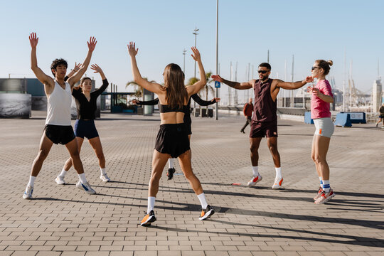 A diverse group of five people exercises outdoors in a circle near a marina, wearing sportswear and performing jumping jacks under the sun. A concept of energy, fitness, and community.