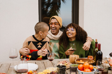 A White bald woman, a White young man with glasses, and a Black young woman laughing and enjoying a lively dinner gathering with wine, fruits, and a warm, cozy atmosphere outdoors.