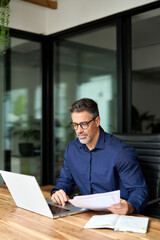 Busy mature male bank manager executive working on laptop checking paper financial report in...