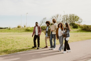 Fototapeta premium Five friends walk along a park pathway, carrying groceries, a camera, and a small dog. They enjoy the fresh air, conversation, and companionship, embracing a relaxed and social outdoor experience.