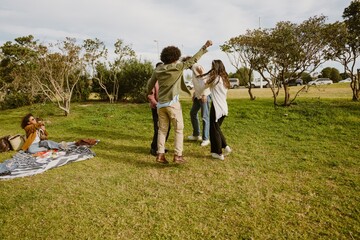 A multinational group of friends having fun, dancing and enjoying a picnic outdoors on a sunny day with a blanket, food, and laughter in the park under the clear blue sky.