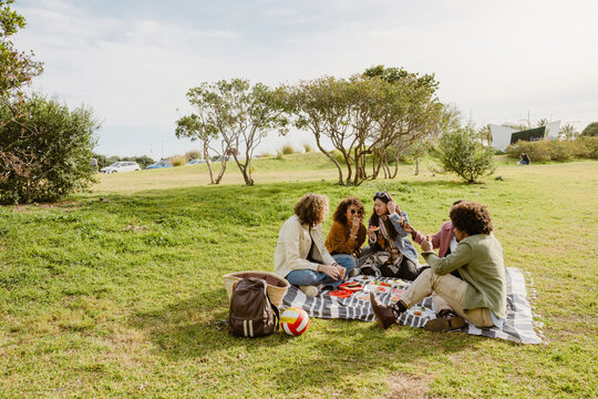 A group of young friends sitting on a blanket in a park, enjoying a picnic. They are eating, laughing, and socializing with nature around them. Various snacks are placed on the picnic setup.