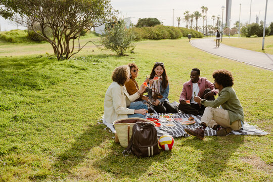 A diverse group of five adults is sitting on a blanket in park, enjoying a picnic with fresh food and watermelon. They are casually dressed, smiling, and talking, surrounded by greenery on sunny day. - Powered by Adobe