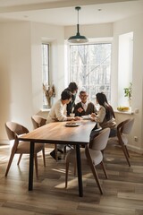 An elderly man sits at a wooden dining table with three younger adults, engaged in a warm conversation. The modern, bright home setting features large windows with natural light.