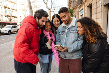 A group of five friends gathers on a city street, looking at a smartphone and smiling. They are dressed in warm jackets, with one person wearing a pink feather boa, suggesting a birthday celebration.