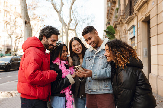 A friends gathers on a city street, laughing and looking at a phone. One person wears a tiara and a pink feather boa, suggesting a birthday celebration or special occasion in a festive atmosphere. - Powered by Adobe