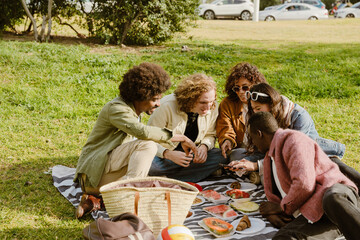 A group of five friends are sitting on a blanket and looking at a phone held by their friend over...