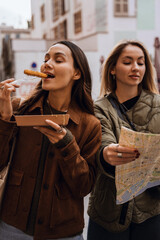 A woman stands with her eyes closed and bites food she holds on a fork, while her friend stands next to her and looks at a map, on the street, while traveling