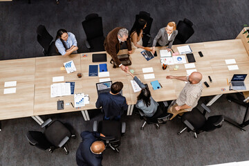 Top view of a group of eight employees standing at a table and taking turns shaking hands, in an office, during a retirement