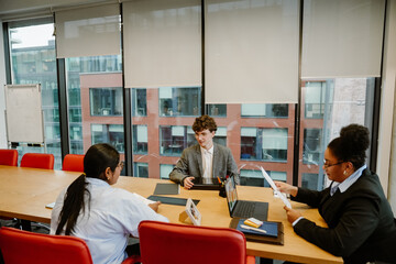 A male intern sits at a table and talks to a female employee sitting across from him, while a...