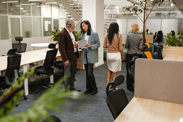 A female employee stands and holds a clipboard while talking to a male employee standing next to...