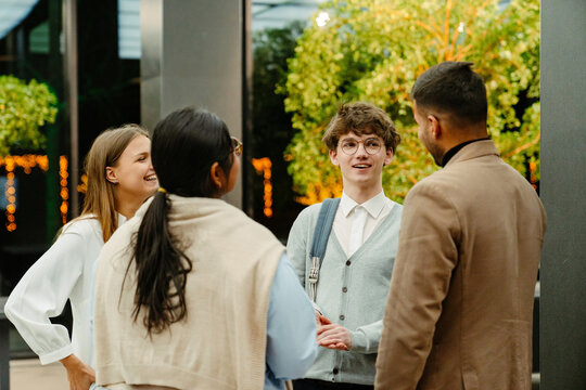 Group of three employees and a male intern stand and laugh while talking and looking at each other, in a corridor, during an internship