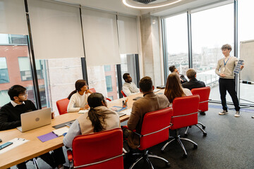 Fototapeta premium A group of eight employees sit at a table and look at a male intern standing next to them and smiling while holding a clipboard, in an office, during an internship