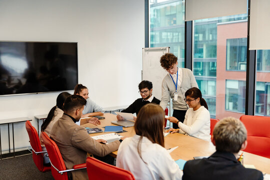 Male intern stands and looks at a table with a group of seven employees sitting at it, in an office, during an internship