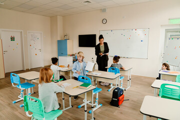The teacher is standing and talking while a student is standing next to her and a group of four students is sitting, at school, in the classroom
