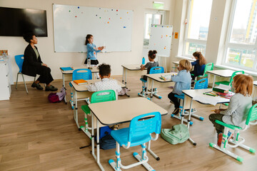 A schoolgirl stands and shows a picture while a teacher and a group of five classmates sit next to her, at school, in the classroom