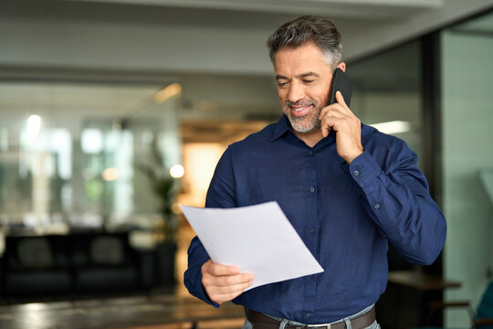 Smiling middle aged senior professional businessman executive looking at documents talking on cell phone standing in office. Happy busy mature older business man holding papers making call at work.