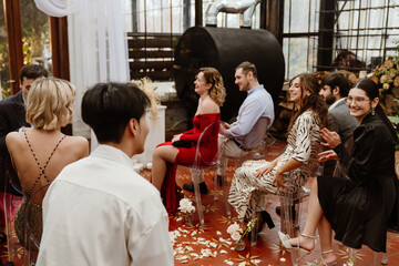 A group of eight guests sit on chairs and smile while talking and looking at each other, before the wedding ceremony