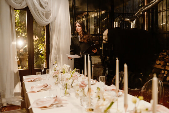 A woman organizer stands and looks at the table while holding a clipboard, in an establishment, before a wedding party