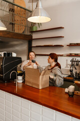 A female employee stands behind the bar and holds a cup next to which the female owner, who stands...