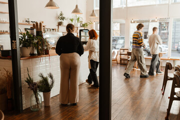Fototapeta premium The woman owner stands and looks at the woman employee who walks by the bar, while two customers pass by them, in the cafe