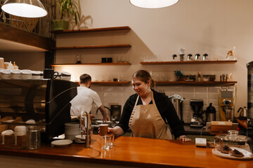 Female owner standing smiling while holding a cup over the bar, male employee standing behind her, in a cafe