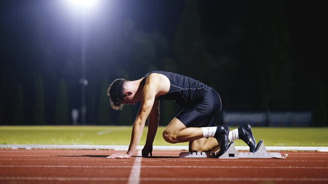 Strong Young Man Starting a Race From Track Starting Blocks Position on a Dark Stadium in the Evening. Cinematic Portrait of a Fit Male Sprint Runner Participating in a Competition