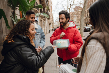 A group of three friends stand and hold packages while looking at a cake held by a friend standing next to them, on their way to a birthday party