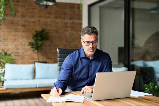 Senior business man company manager working on computer elearning webinar sitting at office desk. Busy mature middle aged professional businessman executive using laptop at work writing notes.